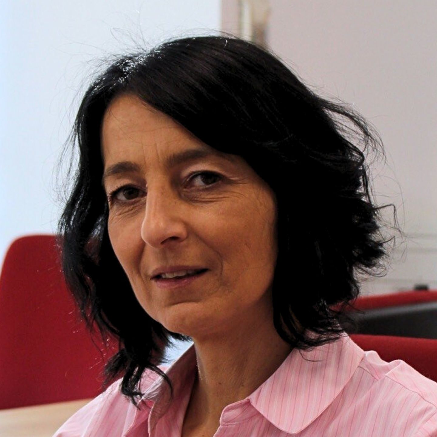Cristina Pasere, az EOS Románia megfelelőségi műveletek támogatásának menedzsere rózsaszín csíkos blúzban ül az asztalánál, és a kamerába néz.