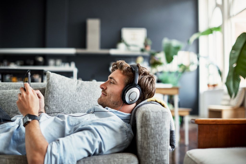 Egy fiatal férfi fekszik a nappalijában egy kanapén, fejhallgatót visel, és mosolyogva nézi az okostelefonját, miközben zenét vagy podcastot hallgat.