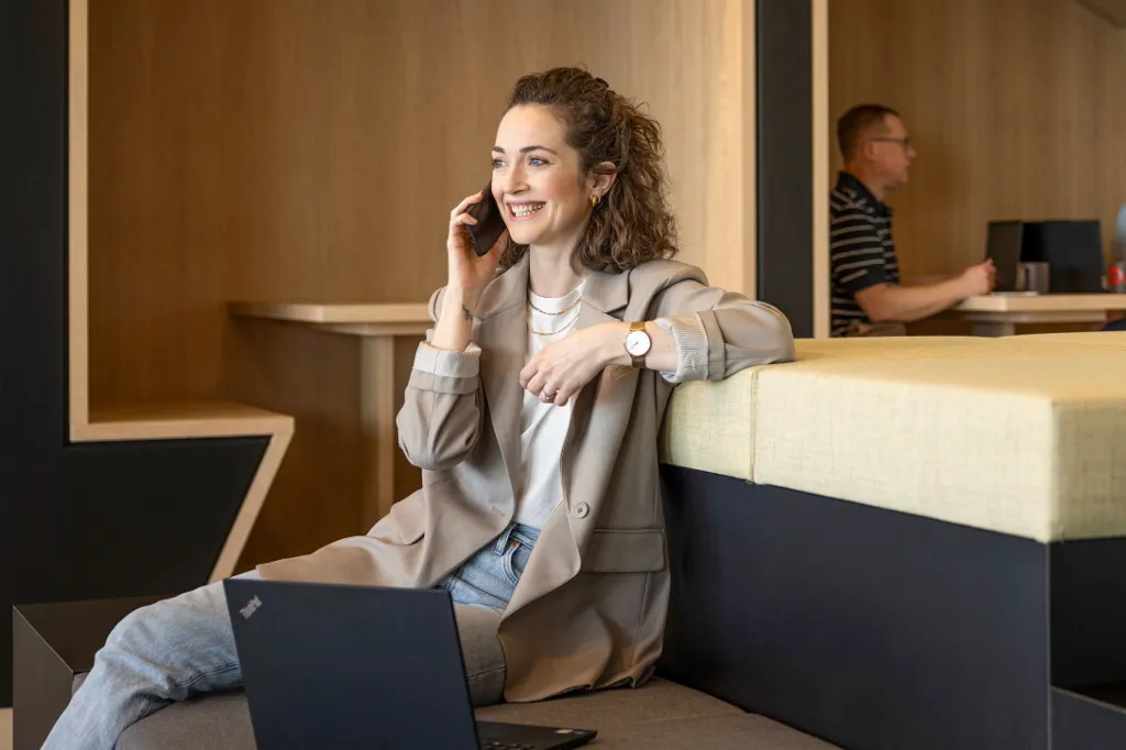 Egy mosolygó személy egy modern irodai lounge-ban ül, telefonon beszél és laptopon dolgozik. A háttérben két másik személy dolgozik.