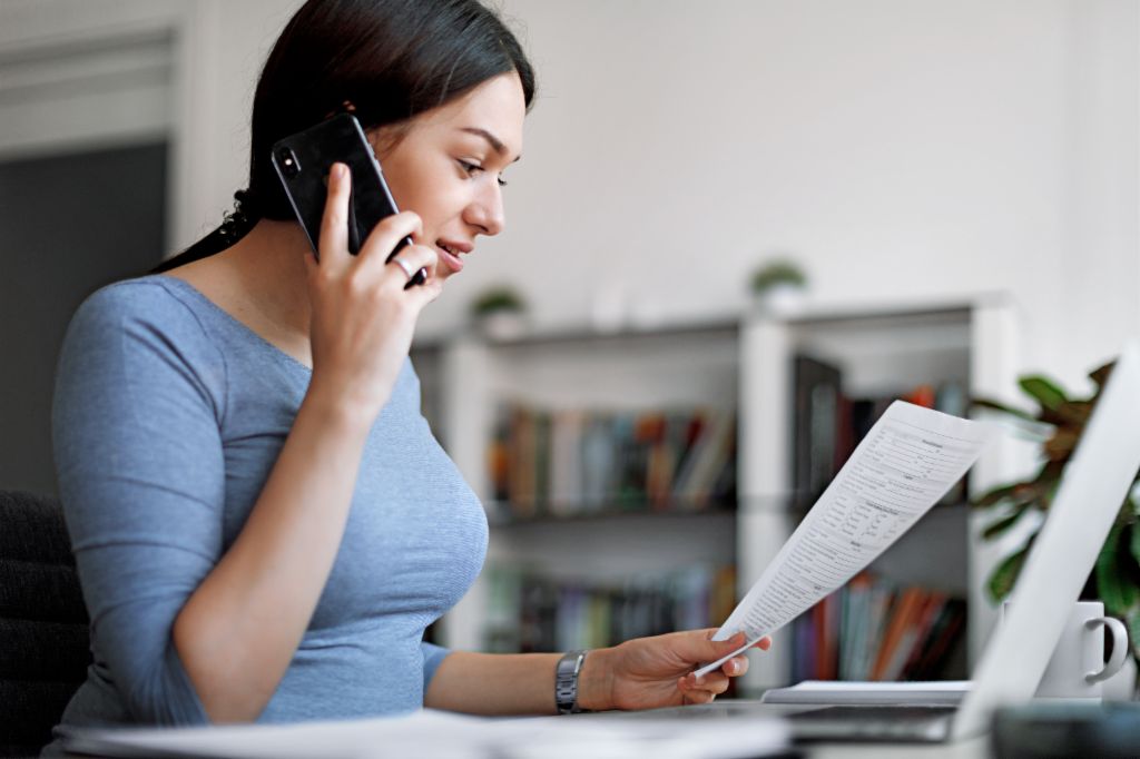 Kék felsőt viselő nő mobiltelefonon beszél miközben a kezében tart egy számlát.