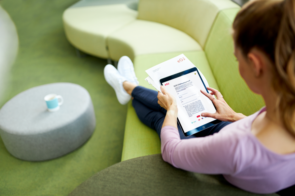 Egy nő ül a kanapén, és egy iPad segítségével fizeti adósságait az EOS Online Portálon.