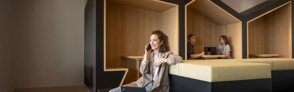 Egy mosolygó személy egy modern irodai lounge-ban ül, telefonon beszél és laptopon dolgozik. A háttérben két másik személy dolgozik.