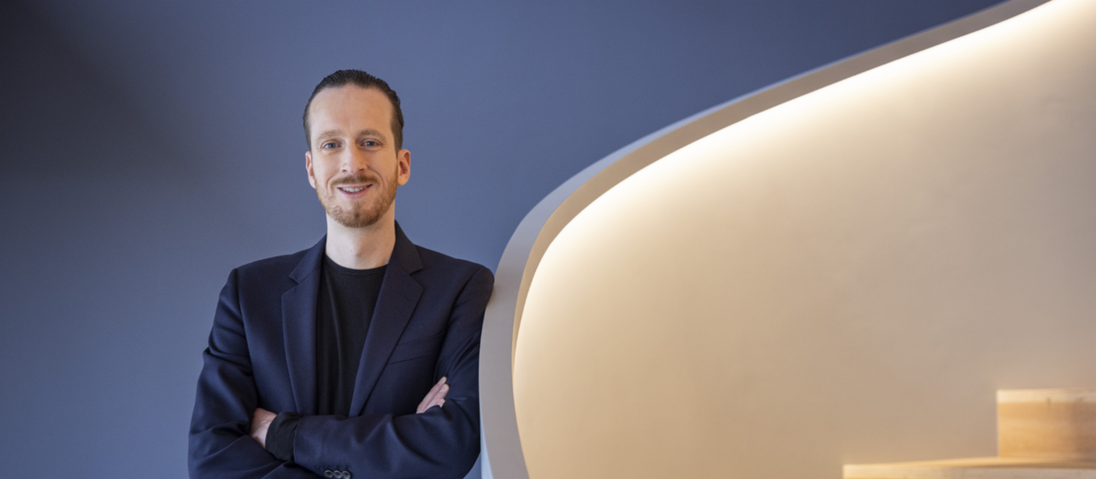 Sebastian Pollmer, egy kék öltönyös férfi, egy falnak támaszkodik egy fehér, meleg fényű lépcső közelében.