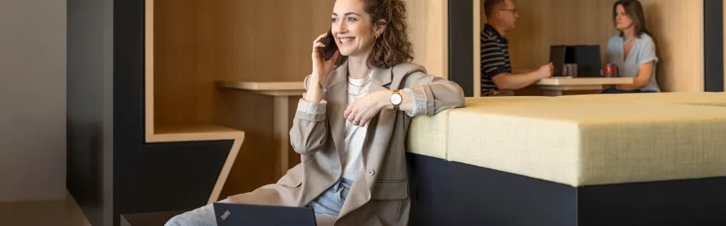 Egy mosolygó személy egy modern irodai lounge-ban ül, telefonon beszél és laptopon dolgozik. A háttérben két másik személy dolgozik.