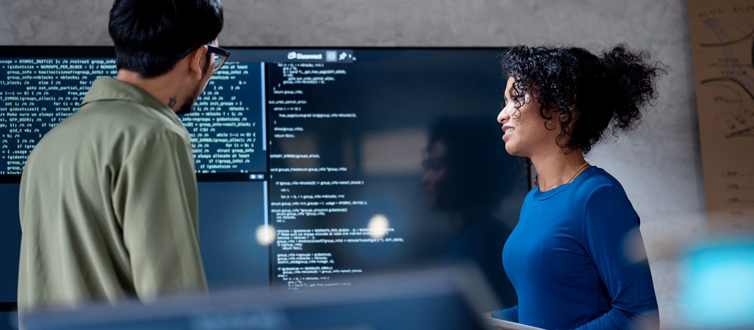 MI a követeléskezelésben: Ezen a képen két szakember közösen elemzi a programkódot automatizált folyamatokhoz egy nagy képernyőn. A jelenet bemutatja a mesterséges intelligencia (MI) és a robotizált folyamatautomatizálás (RPA) gyakorlati alkalmazását a követeléskezelésben – a digitális átalakulás példája a követelések kezelésében.