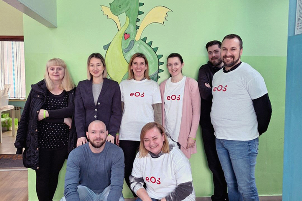 Nyolc ember áll a fal előtt, amelyre egy zöld sárkányt festettek. Az emberek két sorban helyezkednek el: ketten elöl guggolnak, hatan hátul állnak. Négyen piros EOS logóval ellátott fehér pólót viselnek. A csoport belemosolyog a kamerába. A padlón, jobb oldalon elöl néhány palackot és italos kartondobozt látunk.