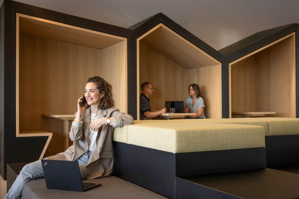 Egy mosolygó személy egy modern irodai lounge-ban ül, telefonon beszél és laptopon dolgozik. A háttérben két másik személy dolgozik.