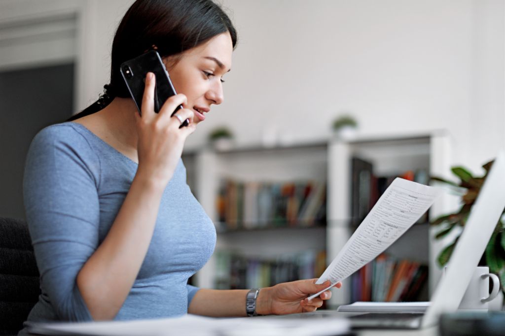 Kék felsőt viselő nő mobiltelefonon beszél miközben a kezében tart egy számlát.